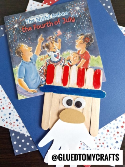 A children’s book titled The Night Before the Fourth of July sits behind a Popsicle Stick Uncle Sam craft, made from popsicle sticks and a paper hand, on star-patterned paper. Text reads @GLUEDTOMYCRAFTS.