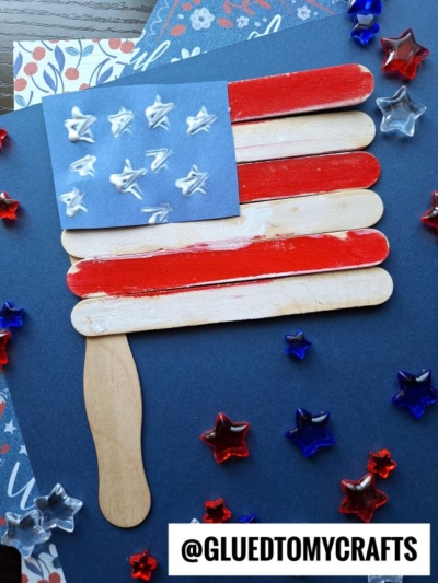 A handmade flag kid craft features colored popsicle sticks and a paper rectangle with puffy white stars, surrounded by red, white, and blue star-shaped beads on a blue background—perfect for creating popsicle stick flags.