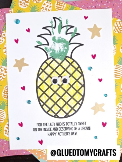 A handmade Mothers Day card features a charming handprint pineapple with googly eyes, surrounded by hearts, stars, and blue sequins. Text below reads: FOR THE LADY WHO IS TOTALLY SWEET ON THE INSIDE AND DESERVING OF A CROWN! HAPPY MOTHERS DAY!.