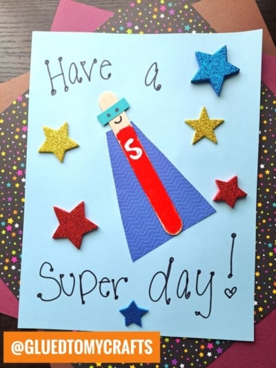 A craft project features the message Have a Super day! with a popsicle stick superhero in a blue mask and red cape, surrounded by colorful glittery stars on a blue paper background. Perfect for fans of popsicle stick superheroes!.