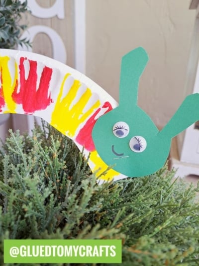 A colorful paper plate caterpillar craft with a green head, googly eyes, and a painted red and yellow body is placed on green foliage. @GLUEDTOMYCRAFTS is written in a green box at the bottom.