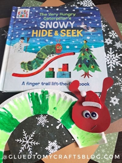 A copy of “The Very Hungry Caterpillar’s Snowy Hide & Seek” is displayed on a snowflake-patterned surface, next to a charming paper plate caterpillar craft with a painted body and red paper face.
