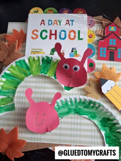 Colorful school-themed craft scene with a book, felt leaves, a paper pencil, a schoolhouse cutout, and two smiling paper plate caterpillars in green and red, arranged on a striped background. @GLUEDTOMYCRAFTS sticker is visible.