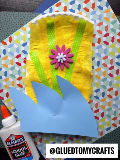 A colorful craft featuring a paper plate surfboard, yellow tissue paper, green polka dot ribbons, a pink flower embellishment, and blue paper cut like grass, all arranged on patterned paper with a bottle of Elmer’s glue. @GLUEDTOMYCRAFTS at the bottom.