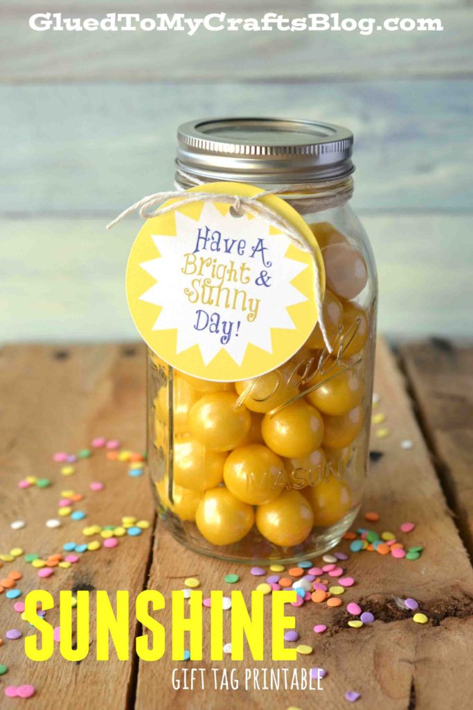 A mason jar filled with yellow candies sits on a wooden surface, topped with a metal lid and a Sunshine Gift Tag reading Have a Bright & Sunny Day! Colorful confetti is scattered around this cheerful sunshine gift idea.