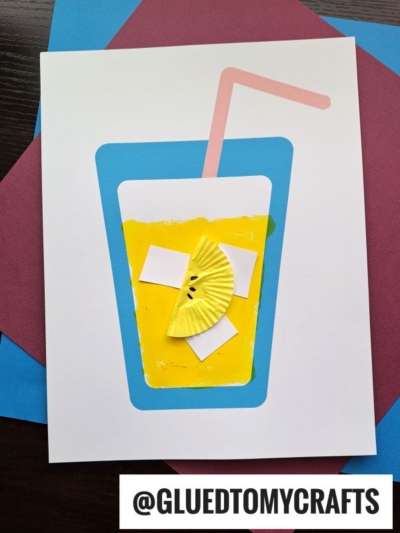 A handmade craft featuring a blue paper cup with cupcake liner lemonade—a yellow drink, pink straw, and a yellow cupcake liner lemon slice with drawn seeds—sits on a white sheet over colored papers. @GLUEDTOMYCRAFTS is at the bottom.