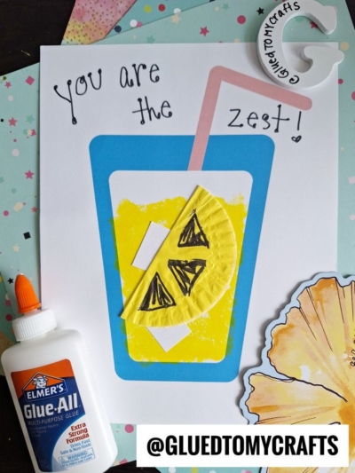 A handmade card features a blue cup of cupcake liner lemonade with a pink straw and a yellow lemon slice. Text reads You are the zest! Nearby are a glue bottle and craft supplies. @GLUEDTOMYCRAFTS is labeled below.