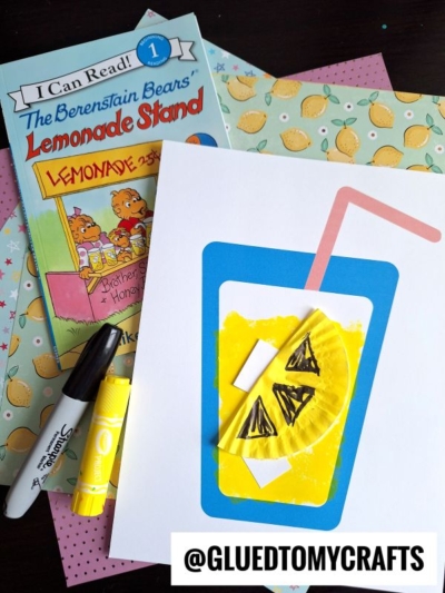 A children’s book, The Berenstain Bears Lemonade Stand, sits next to a cupcake liner lemonade art project on white paper, alongside a marker, glue stick, and patterned papers.