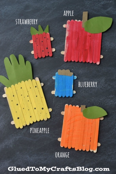 Five colorful popsicle stick fruit crafts—strawberry, apple, blueberry, pineapple, and orange—are arranged on a dark surface, each painted and labeled with its corresponding fruit name.