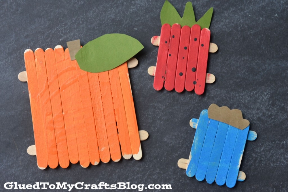 Three popsicle stick fruit shapes: an orange pumpkin with a green leaf, a red strawberry with green leaves and black dots, and a blue rectangle with a brown top, all crafted from colored popsicle sticks on a dark background.