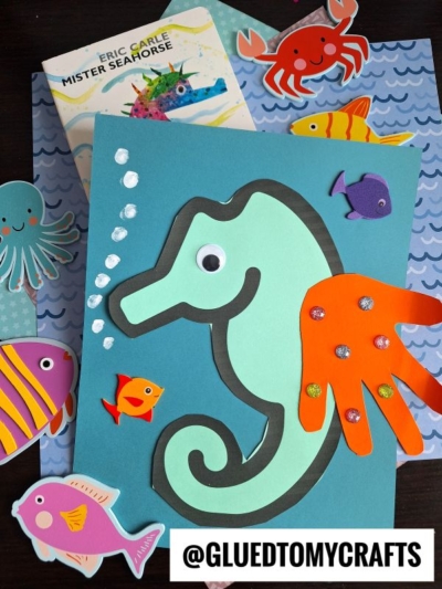 A seahorse craft made with cut paper and a child’s handprint is surrounded by colorful paper sea creatures, set on blue patterned paper—perfect for Eric Carle crafts—with the book Mister Seahorse by Eric Carle in the background.