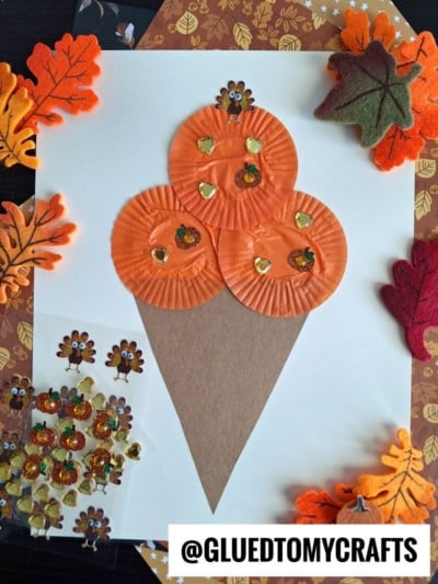A fall-themed ice cream kid craft shaped like a cone, made with three orange cupcake liners and a brown paper cone, decorated with turkey stickers, gold sequins, and surrounded by felt leaves and autumn-patterned paper.