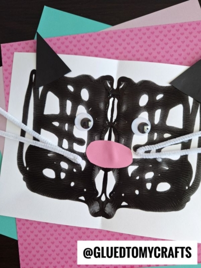 A symmetrical black cat face made from paint, with googly eyes, a pink paper nose, white pipe cleaner whiskers, and black paper ears—a fun Eric Carle craft displayed on layered colored paper.