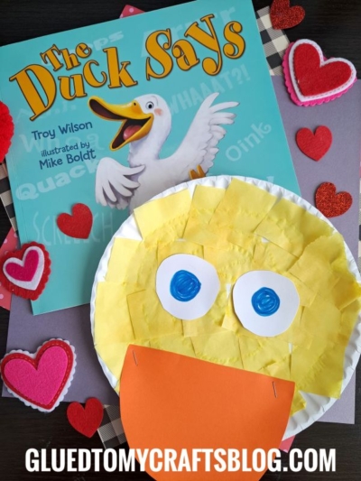 A children’s book titled The Duck Says sits beside a duck face craft made with a paper plate, yellow squares, and orange paper—perfect inspiration for story time crafts. Red felt hearts and GLUEDTOMYCRAFTSBLOG.COM decorate the background.