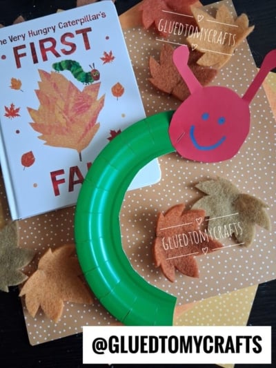 A children’s autumn craft features a charming paper plate caterpillar and a book titled The Very Hungry Caterpillar’s First Fall, surrounded by cut-out felt leaves on a polka-dot surface.