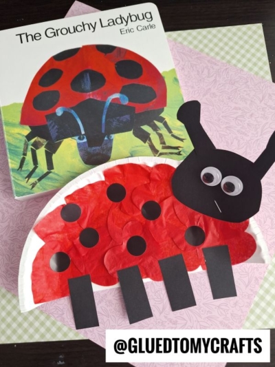 A copy of The Grouchy Ladybug by Eric Carle sits beside a creative Eric Carle craft—a ladybug made from a paper plate, tissue paper, and googly eyes—perfect for story time crafts! @GLUEDTOMYCRAFTS.