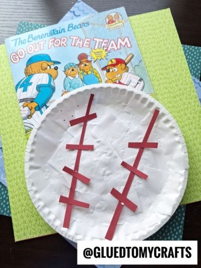 A Berenstain Bears book titled Go Out for the Team sits behind a paper plate baseball craft, with red paper strips glued on as stitching. The background includes patterned green and blue paper.