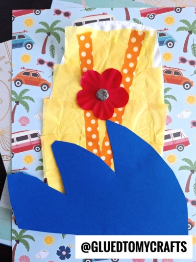 A paper craft of a shark fin in blue paper, set on a yellow background with orange stripes, a red flower, and a paper plate surfboard, all on beach-themed paper featuring cars and palm trees. The handle @GLUETOMYCRAFTS is at the bottom.