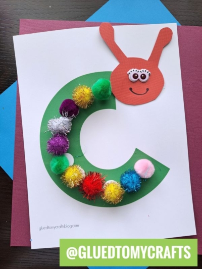 A fun Eric Carle craft project featuring a large green letter C decorated with colorful pom-poms, topped with a red paper snail head with googly eyes and a smile, perfect for story time crafts on a white sheet of paper.