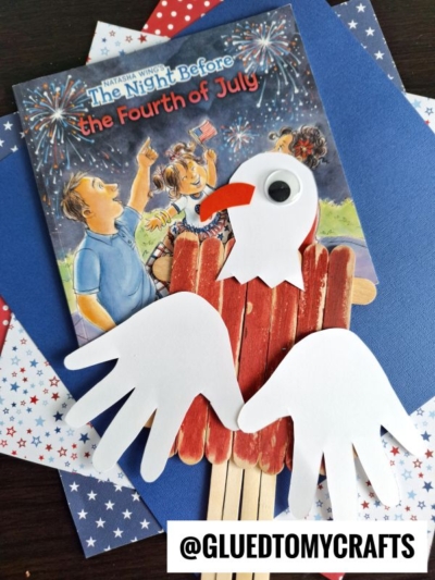 A children’s book titled The Night Before the Fourth of July lies on star-patterned paper, beside a craft stick bald eagle with paper wings and a googly eye. @GLUEDTOMYCRAFTS is written at the bottom.