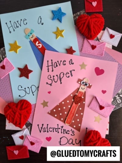 Two handmade Valentine’s Day cards decorated with stars, hearts, and popsicle stick superheroes using craft sticks and paper. Surrounding the cards are red yarn hearts and mini felt envelopes. @GLUEDTOMYCRAFTS appears at the bottom.