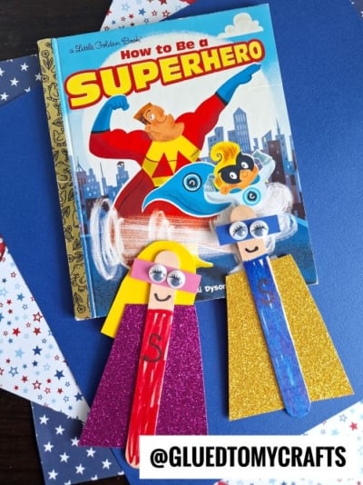 Two colorful popsicle stick superheroes made from googly eyes, paper capes, and felt masks are displayed on a star-patterned surface next to the book How to Be a Superhero. @GLUEDTOMYCRAFTS is written at the bottom.