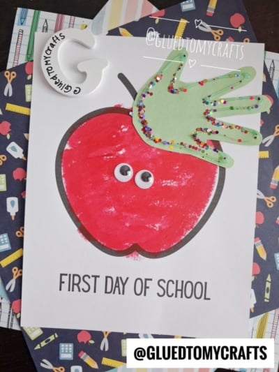 Handprint craft of a red apple with googly eyes and a green paper hand decorated with sequins on top, labeled First Day of School—perfect as an adorable apple handprint keepsake. A ceramic paint palette with GLUEDTOMYCRAFTS is in the top left.