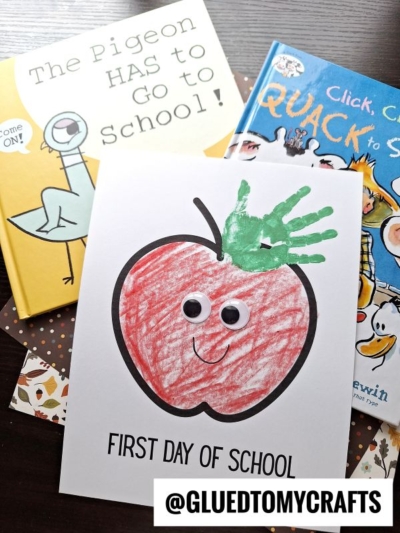 Childrens books and a First Day of School craft featuring an adorable red apple handprint keepsake with googly eyes, a smiley face, and a green handprint as the leaf. The handle @GLUEDTOMYCRAFTS is displayed.