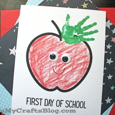 A handmade card with a large red crayon apple, googly eyes, a green apple handprint keepsake as the leaf, a smiley face, and “FIRST DAY OF SCHOOL.” The background features patterned paper