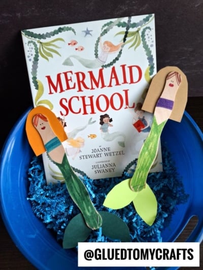 A children’s book titled Mermaid School stands in a blue bucket with shredded blue paper, accompanied by two handmade craft stick mermaid creations made from popsicle sticks, paper, and markers. A tag reads @GLUEDTOMYCRAFTS.