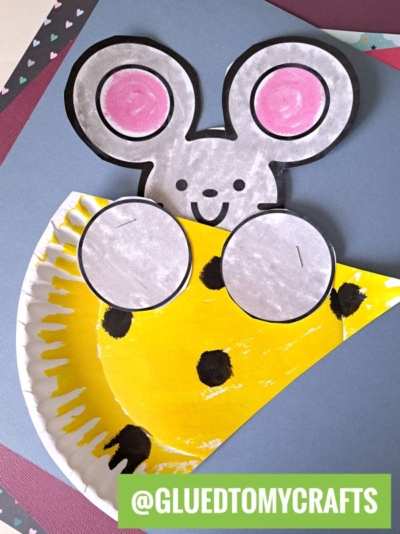 A paper craft features a gray mouse with pink ears peeking over a yellow, paper plate cheese with black spots. The image includes the text @gluedtomycrafts at the bottom.