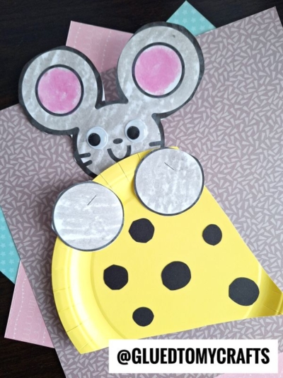 A paper craft mouse with large pink ears peeks out from behind a yellow paper plate cheese wedge. Background includes pink and blue patterned papers. Text reads @GLUEDTOMYCRAFTS.