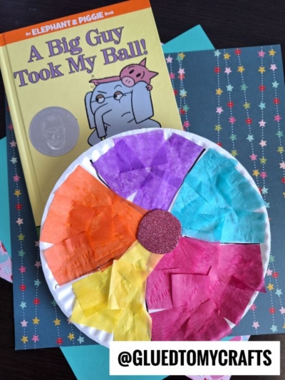 A children’s book titled A Big Guy Took My Ball! lies on patterned paper next to a vibrant paper plate beach ball craft, featuring sections of orange, purple, blue, yellow, and pink tissue paper. @GLUEDTOMYCRAFTS watermark is in the bottom corner.