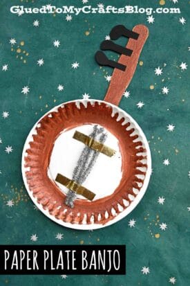 A homemade paper plate banjo craft painted brown, with a cardboard neck and black tuning pegs, decorated with gold tape and silver pipe cleaners. Set on a green background with white stars. Text: Paper Plate Banjo.