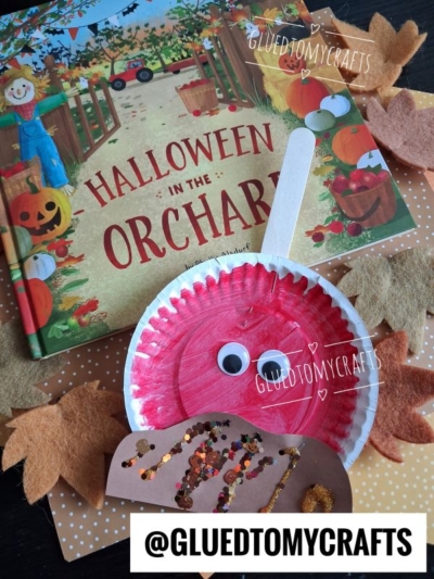 A Halloween in the Orchard book, a red paper plate apple craft with googly eyes, and a popsicle stick—reminiscent of festive paper plate candy apples—are surrounded by paper leaves and a glittery paper pie. @GLUEDTOMYCRAFTS watermark is visible.