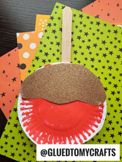 A paper plate painted red with brown glitter paper on top and a popsicle stick handle, resembling one of those classic paper plate candy apples, sits on Halloween-themed paper. @GLUEDTOMYCRAFTS is labeled at the bottom.