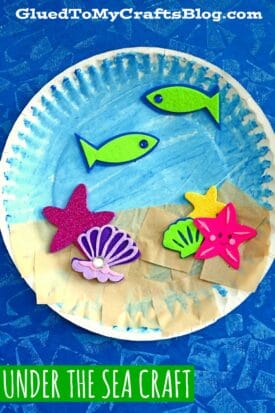 A paper plate ocean painted blue features foam sea creatures—fish, starfish, and shells—plus tan paper strips for sand, creating a vibrant under the sea craft scene. Text reads, UNDER THE SEA CRAFT.
