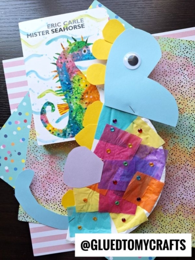 A colorful tissue paper seahorse craft made from paper, tissue, and sequins sits on printed papers beside the book Mister Seahorse by Eric Carle. The seahorse features a googly eye and bursts with bright colors.