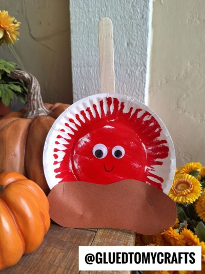 A paper plate craft of a red hedgehog with googly eyes, a brown paper base, and a wooden stick handle—reminiscent of paper plate candy apples—is surrounded by pumpkins and yellow flowers.
