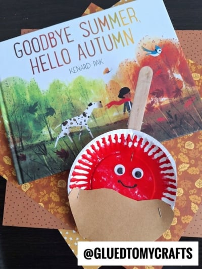 A children’s book titled Goodbye Summer, Hello Autumn by Kenard Pak sits on patterned paper, next to a red paper plate candy apple craft with googly eyes, a smile, popsicle stick stem, and tan paper leaf.