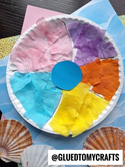 A paper plate beach ball craft divided into colored sections with tissue paper in pink, purple, orange, yellow, and blue, a blue center circle, seashells, and a sign reading @GLUEDTOMYCRAFTS nearby.