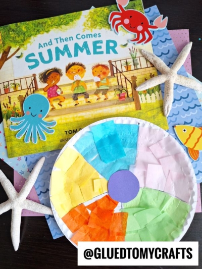 Children’s book And Then Comes Summer with a colorful beach ball paper plate craft, sea-themed decorations, starfish, and ocean animal cutouts on a table. ©GLUEDTOMYCRAFTS.