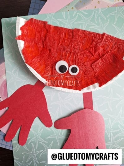 A paper plate red crab craft for kids, made with a painted plate, googly eyes, and red paper claws and legs, sits on patterned paper. The watermark @GLUEDTOMYCRAFTS is visible at the bottom.