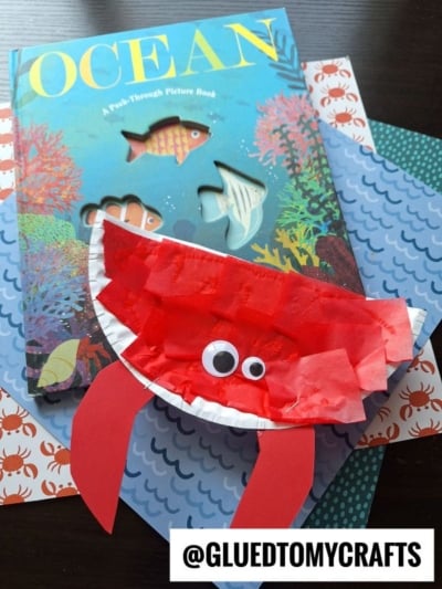 A children’s ocean-themed book with colorful fish on the cover sits on blue and crab-patterned paper, next to a playful paper plate red crab craft with big googly eyes and red paper claws.