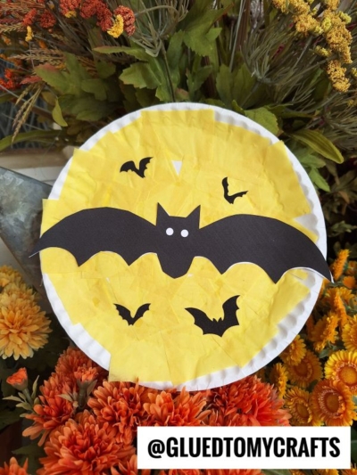 A paper plate yellow moon with black bat cutouts is displayed among orange and yellow flowers. The handle @GLUEDTOMYCRAFTS appears at the bottom of the image.