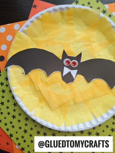 A paper plate yellow moon is decorated with bright yellow tissue paper, while a black paper bat with striking red eyes is glued on top. Festive, Halloween-themed paper sits underneath to complete this playful paper plate fright night craft.