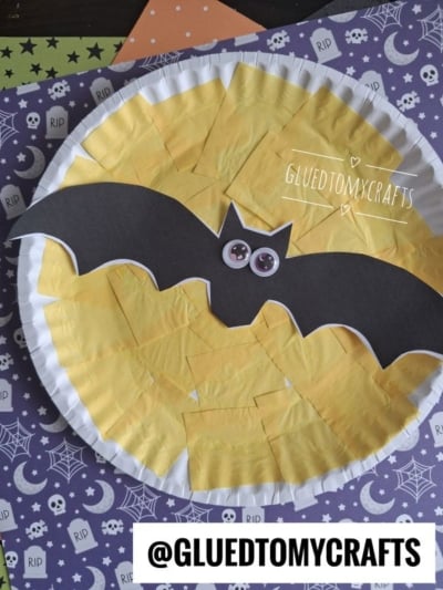 A paper plate yellow moon made with yellow tissue squares forms a glowing lunar shape, topped with a black bat cutout and googly eyes, all set on Halloween-themed paper for the perfect paper plate fright night craft.
