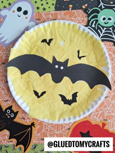 A paper plate yellow moon with yellow tissue paper, black paper bats, and Halloween-themed decorations like a ghost, spider, and pumpkin in the background. @GLUEDTOMYCRAFTS is written at the bottom.