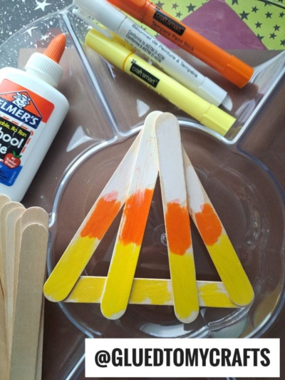 Wooden craft sticks painted yellow, orange, and white are arranged in a triangle on a tray, resembling a popsicle stick candy corn. Nearby are glue, orange and yellow markers, and unpainted sticks. Text reads @GLUEDTOMYCRAFTS.