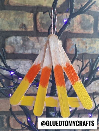 Three wooden craft sticks painted yellow and orange are glued in a triangular shape, resembling a popsicle stick candy corn campfire, and hanging from a black tree branch. Background is a brick wall. The text @GLUEDTOMYCRAFTS is at the bottom.